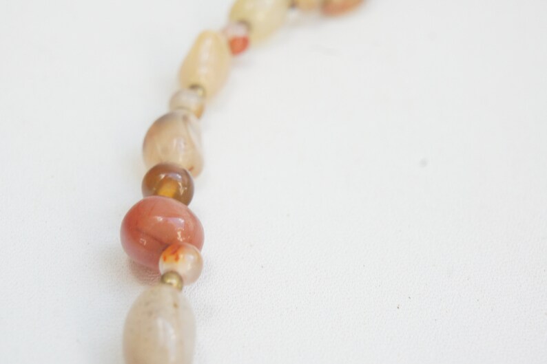 May include: A close-up of a necklace made of natural stone beads. The beads are various shades of brown, orange, and white. The necklace is strung on a thin cord.