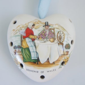May include: A heart-shaped ceramic ornament with a light blue ribbon hanger. The front features a colorful illustration of two people in traditional attire at a table, with the text SOUVENIR OF WALES printed below. The edges have small circular holes.