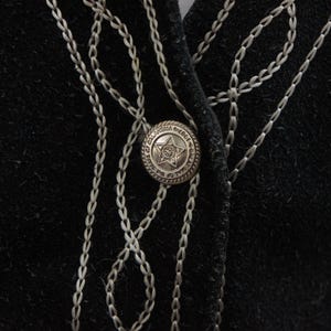 May include: Close-up of a black garment with silver chain-like embroidery and a decorative button. The button is round with a star design in the center and a detailed border. The embroidery forms a pattern around the button and along the garment's edges.