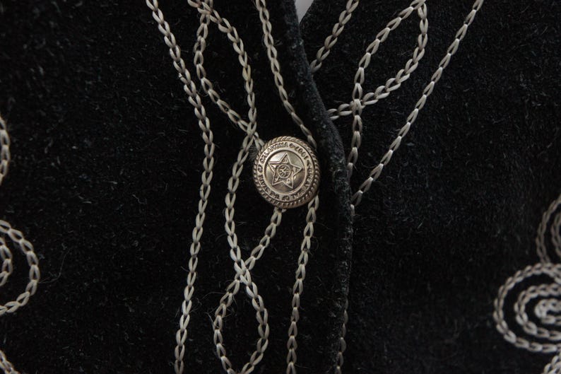 May include: Close-up of a black garment with silver chain-like embroidery and a decorative button. The button is round with a star design in the center and a detailed border. The embroidery forms a pattern around the button and along the garment's edges.