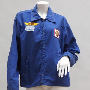 May include: A blue zip-up jacket with a collared neckline and two front pockets. The jacket features patches on the chest, including a yellow and white patch with the word "Beechcraft". The jacket is displayed on a mannequin.