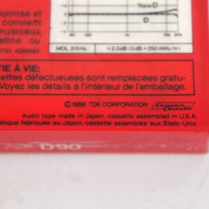 Puede incluir: Primer plano de una caja de cinta de casete roja con el texto "TDK D90" impreso en la parte delantera. La parte posterior de la caja tiene una declaraci&oacute;n de garant&iacute;a en franc&eacute;s e ingl&eacute;s, y el texto "&copy;1986 TDK CORPORATION" impreso en negro.