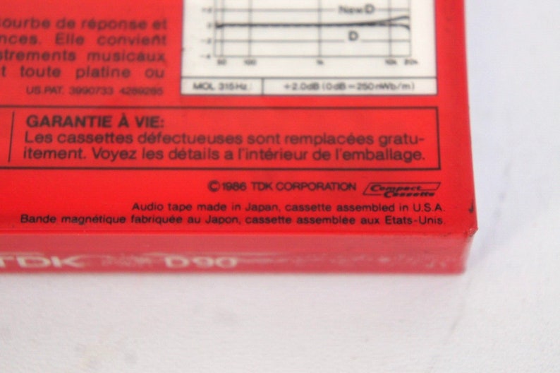 Puede incluir: Primer plano de una caja de cinta de casete roja con el texto "TDK D90" impreso en la parte delantera. La parte posterior de la caja tiene una declaraci&oacute;n de garant&iacute;a en franc&eacute;s e ingl&eacute;s, y el texto "&copy;1986 TDK CORPORATION" impreso en negro.