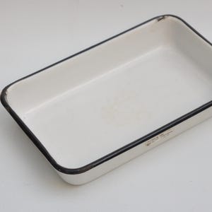 Op de afbeelding: Rechthoekige witte emaille bakvorm met zwarte rand. De vintage vorm heeft een licht versleten uiterlijk, wat duidt op leeftijd en gebruik. De vorm is leeg en ligt op een wit oppervlak.