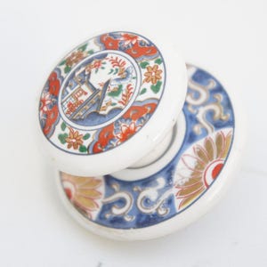 May include: Two white ceramic door knobs with a decorative design. The knobs feature a circular pattern with red, orange, blue, and green floral and architectural motifs. The knobs are stacked on top of each other.