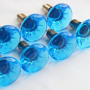 May include: A set of seven cabinet knobs featuring translucent, bright blue glass tops with a fluted, petal-like design and a metallic, brass-toned cylindrical base. Each knob has a smooth, rounded surface and a distinct, radiating pattern.