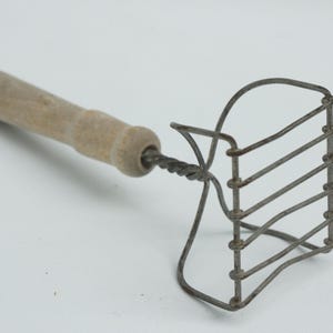May include: A vintage potato masher with a light-colored wooden handle and a wire head. The wire head is made of several parallel horizontal wires connected by vertical wires. The handle is attached to the head with a twisted wire.