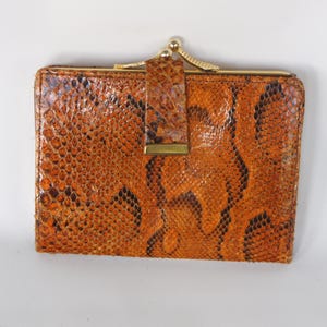 May include: A rectangular bi-fold wallet featuring a textured brown and orange snakeskin pattern. The wallet includes a gold-toned metal kiss-lock clasp at the top and a matching vertical strap with a gold-toned metal accent on the front exterior.