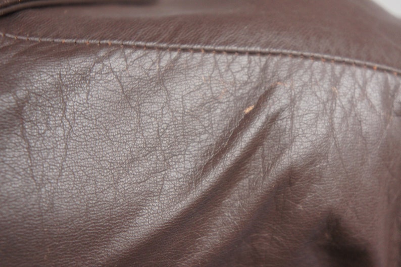 May include: Close-up of a brown leather jacket. The leather has a textured, wrinkled appearance with visible stitching along the seams. The color is a rich, dark brown.