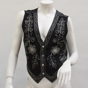 May include: Black vest with silver floral embroidery. The vest features a V-neck, button closure, and decorative stitching along the edges. The vest is displayed on a mannequin.