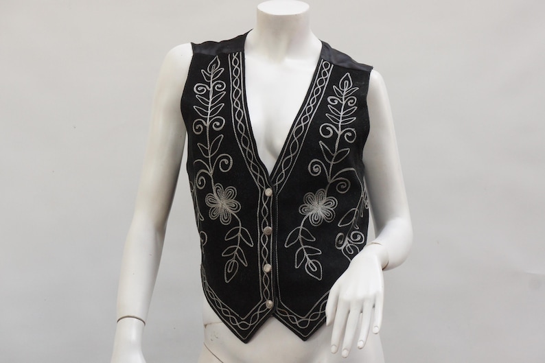 May include: Black vest with silver floral embroidery. The vest features a V-neck, button closure, and decorative stitching along the edges. The vest is displayed on a mannequin.