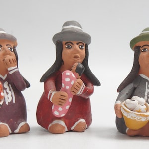 Könnte beinhalten: Drei handgefertigte Keramikfiguren, die Frauen in traditioneller Kleidung darstellen. Jede Figur trägt einen Hut und hält Gegenstände wie ein Baby, Obst oder Essenskörbe. Die Figuren sind in Erdtönen mit Akzenten in Rot, Weiß und Grau bemalt.