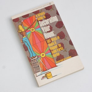 以下が含まれることがあります： アン・パークス・バーク著『A Groovy Guide to Decorating Your Room: 501 Ways to Make It Happen』というタイトルのヴィンテージペーパーバック本。表紙には、脚立に乗って大きくカラフルな幾何学模様を描いている人物のイラストが描かれています。