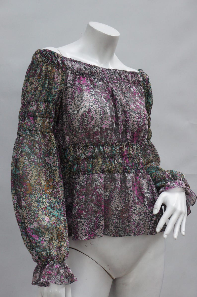 May include: A floral print off-the-shoulder blouse. The sheer fabric features a pattern of small flowers in shades of pink, green, and purple on a dark background. The blouse has long sleeves with ruffled cuffs and a gathered waist.