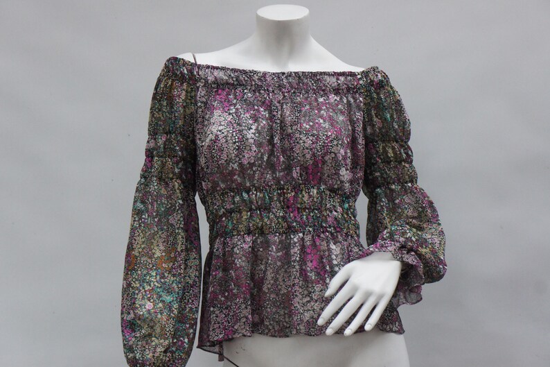 May include: Off-the-shoulder blouse with long sleeves. The sheer fabric features a floral pattern in shades of purple, green, and brown. The blouse has elastic at the neckline and waist, and is displayed on a mannequin.