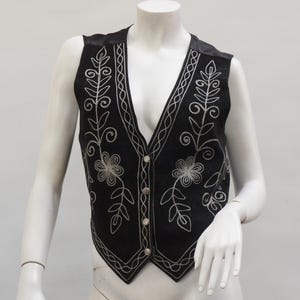 May include: Black vest with silver floral embroidery. The vest has a V-neck, button closure, and decorative stitching. The garment is displayed on a mannequin.