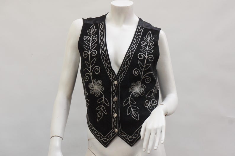 May include: Black vest with silver floral embroidery. The vest features a V-neck, button closure, and decorative stitching along the edges. The vest is displayed on a mannequin.