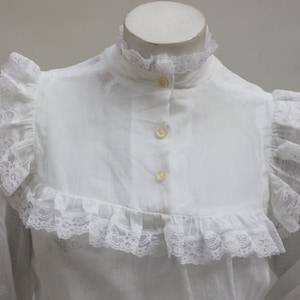 May include: White blouse with a high lace collar and lace trim around the neckline and sleeves. The blouse has a button-up front.