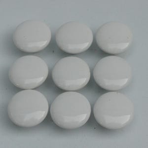 Pode incluir: Nove puxadores de armário redondos de cerâmica branca dispostos em padrão de grade. Cada puxador tem um acabamento brilhante, refletindo a luz. Estes puxadores são adequados para gavetas, armários ou outros móveis.