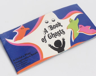 以下が含まれることがあります： 「A Book of Ghosts」というタイトルのヴィンテージ子供向けの本。表紙は青色の背景に、オレンジ、ピンク、白の幽霊のイラストが描かれています。本のタイトルは黒い文字で、著者の名前も表示されています。