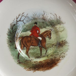 Puede incluir: Un platillo de cer&aacute;mica blanca con un borde rosa presenta una escena pintada a mano de un jinete a caballo en un entorno boscoso. El jinete lleva una chaqueta roja y pantalones de montar negros. El caballo es marr&oacute;n con una estrella blanca en la cara.