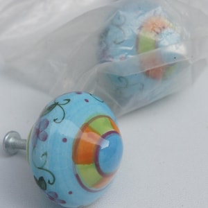 May include: Blue ceramic cabinet knobs with a colorful design. The knobs have a light blue base with orange, green, and purple accents. The knobs have a silver metal screw for installation. One knob is out of the packaging.