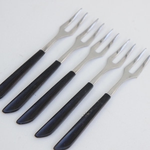 Könnte beinhalten: Sechs Vintage-Gabeln aus Edelstahl mit schwarzen Griffen. Die Gabeln haben ein klassisches Design mit vier Zinken und sind diagonal auf einer weißen Oberfläche angeordnet. Die Griffe sind einfarbig schwarz.