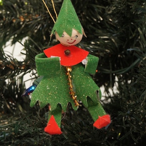May include: A Christmas tree ornament shaped like an elf with a green felt hat, a red collar, and a green felt body with gold glitter accents. The elf has a painted wooden face, red pointed shoes, and a gold tinsel strip down the center.