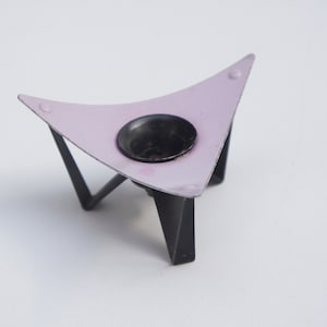 May include: A pink metal candle holder with a black metal base. The holder is shaped like a triangle with a round opening in the center.