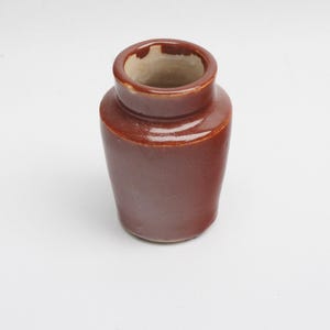 May include: A small, brown ceramic jar with a cylindrical body and a slightly flared rim. The jar has a glossy finish and a simple, classic design. The interior is a lighter tan color.