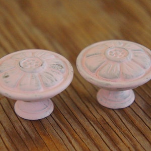May include: Two pink flower-shaped cabinet knobs with a distressed white finish.