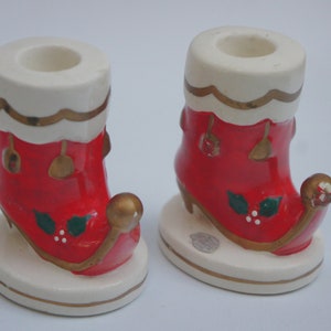 May include: Pair of ceramic Christmas stocking candle holders. The red stockings have white trim with gold accents and holly details. The candle holders are on a white base with a gold trim.