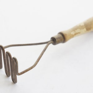 May include: A vintage potato masher with a wooden handle and a metal mashing head. The handle is light brown with signs of wear, and the metal head has a wavy design. This kitchen utensil is designed for mashing potatoes and other soft foods.