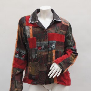 May include: A patchwork jacket with a button-up front, displayed on a mannequin. The jacket features a mix of textures and colors, including red corduroy, embroidered details, and various patterned fabrics. The collar is dark with white stitching.