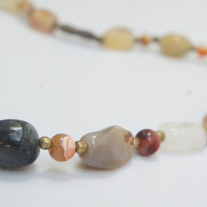 May include: A necklace made of various natural stones, including grey, white, brown, and red. The stones are strung on a brown cord.
