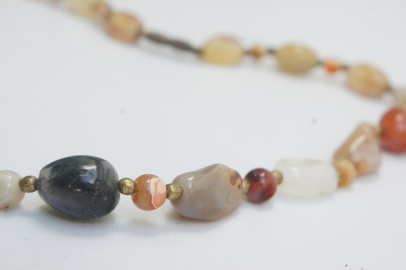 May include: A necklace made of various natural stones, including grey, white, brown, and red. The stones are strung on a brown cord.