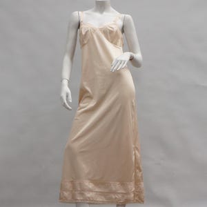 Op de afbeelding: Een perzikroze slipdress met spaghettibandjes en een kanten rand langs de halslijn en zoom. De jurk wordt gepresenteerd op een mannequin tegen een effen achtergrond. De slipdress is een vintage stijl.
