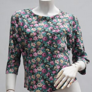Puede incluir: Una blusa de estampado floral con fondo oscuro y flores rosas, blancas y verdes. La blusa tiene un bolsillo en el lado izquierdo y mangas tres cuartos.