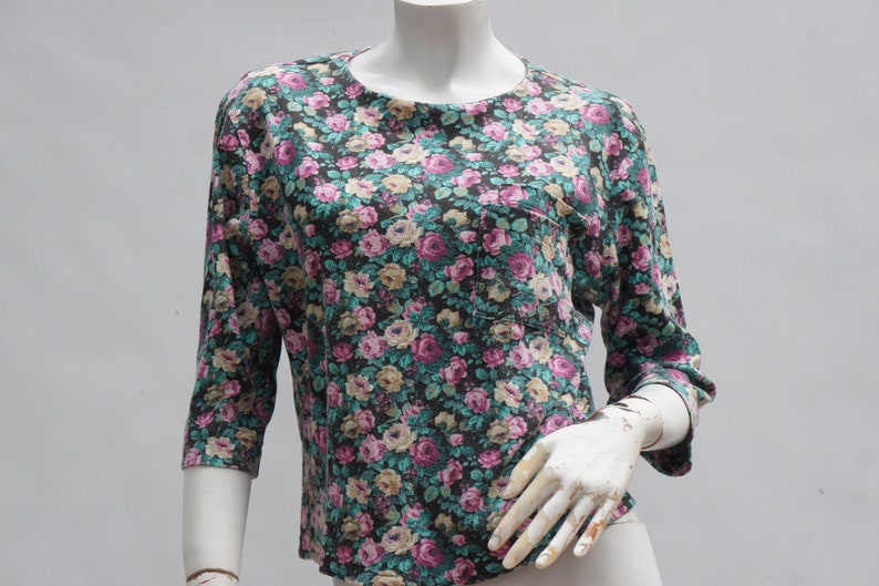 Puede incluir: Una blusa de estampado floral con fondo oscuro y flores rosas, blancas y verdes. La blusa tiene un bolsillo en el lado izquierdo y mangas tres cuartos.