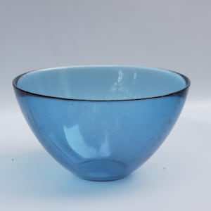 May include: A blue glass bowl with a rounded shape and a dark rim. The bowl has a light blue interior and a smooth, glossy exterior. The bowl is empty and sits on a white surface.