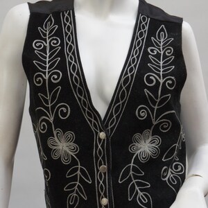 May include: Black vest with silver floral embroidery. The vest features a V-neck, button closure, and decorative stitching along the edges. The embroidery includes flowers and leaf patterns. The vest is displayed on a mannequin.