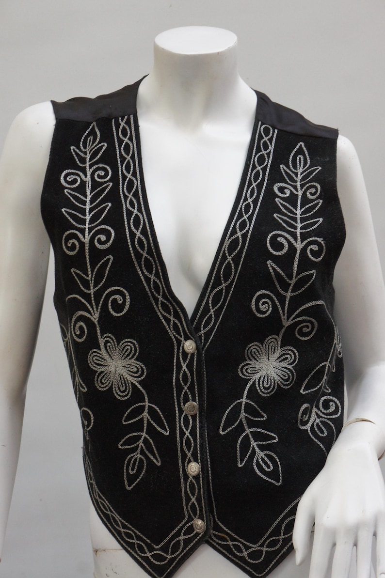 May include: Black vest with silver floral embroidery. The vest features a V-neck, button closure, and decorative stitching along the edges. The embroidery includes flowers and leaf patterns. The vest is displayed on a mannequin.