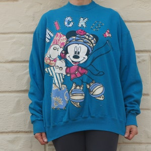 May include: A bright blue mock neck sweatshirt featuring a large graphic of Mickey Mouse wearing a hat and scarf. The design includes patchwork-style elements with various patterns and the word MICKEY spelled out in stitched-look letters.
