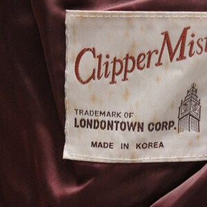 May include: Close-up of a label with the text "ClipperMist" in a reddish-brown script. The label also includes the text "TRADEMARK OF LONDON TOWN CORP. MADE IN KOREA" and a graphic of a building. The background is a dark burgundy fabric.