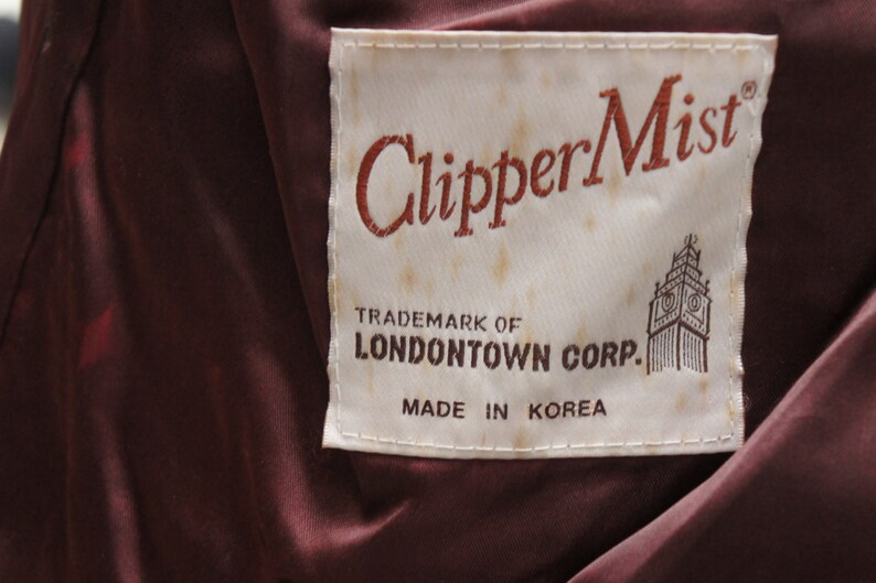 May include: Close-up of a label with the text "ClipperMist" in a reddish-brown script. The label also includes the text "TRADEMARK OF LONDON TOWN CORP. MADE IN KOREA" and a graphic of a building. The background is a dark burgundy fabric.