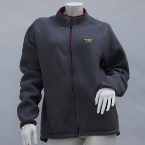 May include: A charcoal gray fleece jacket with a full zipper and a stand-up collar. The jacket has burgundy trim on the collar, cuffs, and zipper. A small rectangular patch is on the left chest.