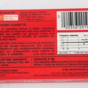 Puede incluir: Caja roja de cinta de cassette con un c&oacute;digo de barras, informaci&oacute;n del producto y un gr&aacute;fico de respuesta de frecuencia. La caja est&aacute; etiquetada como "TDK D90 Cassette Din&aacute;mico" y ofrece una garant&iacute;a de por vida.