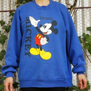 以下が含まれることがあります： MICKEYとNEW MEXICOの文字がプリントされた、ロイヤルブルーの長袖スウェットシャツ。黒、白、赤、黄色の色使いで描かれたミッキーマウスのグラフィックが、青い衣類の前面に配置されています。