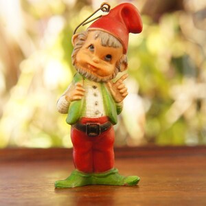 May include: A vintage gnome figurine with a red hat, green vest, and red pants. The gnome has a white shirt, a black belt, and green shoes. The figurine is standing and pointing with one finger. The background is blurred.