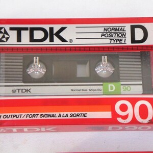 Puede incluir: Una caja de cassette de audio roja, blanca y negra con el texto "TDK" y "D 90" impreso en ella. La caja est&aacute; etiquetada "Posici&oacute;n Normal Tipo I" y "Alta Salida/Se&ntilde;al Fuerte a la Salida".
